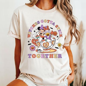 K&ouml;nnte beinhalten: Ein beiges T-Shirt mit einem Aufdruck von Mickey Mouse und Daisy Duck, die als Hexen in einer Teetasse verkleidet sind. Der Text "Witches Gotta Stick Together" ist auf dem Shirt aufgedruckt.