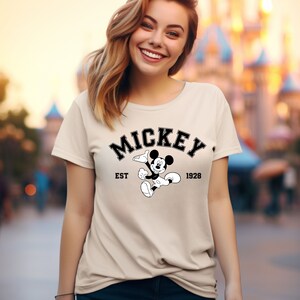 May include: A beige t-shirt with a black and white graphic of Mickey Mouse. The graphic includes the text "Mickey" in a black arch above Mickey Mouse, "EST" to the left of Mickey Mouse, and "1928" to the right of Mickey Mouse.