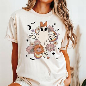 May include: A white t-shirt with a cartoon ghost wearing a pink bow and surrounded by flowers and pumpkins. The ghost has large eyes and a surprised expression. The design is in a retro style with rainbow accents.