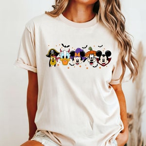 Op de afbeelding: Een beige T-shirt met een grafische print van Mickey Mouse, Donald Duck, Goofy en Pluto in Halloween-kostuums. De personages staan in een rij met vleermuizen en oranje stippen op de achtergrond.