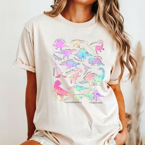 May include: A light beige t-shirt with a graphic print of various dinosaurs in rainbow colors. The dinosaur names are listed below each dinosaur silhouette.