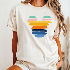 May include: A cream colored t-shirt with a colorful Mickey Mouse silhouette design. The silhouette is made up of horizontal stripes in shades of green, pink, orange, yellow, blue, and light blue.