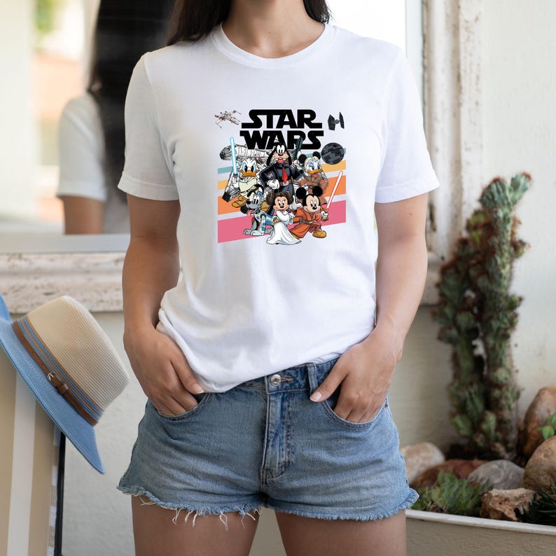 K&ouml;nnte beinhalten: Wei&szlig;es T-Shirt mit einer farbenfrohen Grafik von Disney-Charakteren in Star Wars-Kleidung. Das Star Wars-Logo ist &uuml;ber den Charakteren aufgedruckt. Das T-Shirt wird getragen.