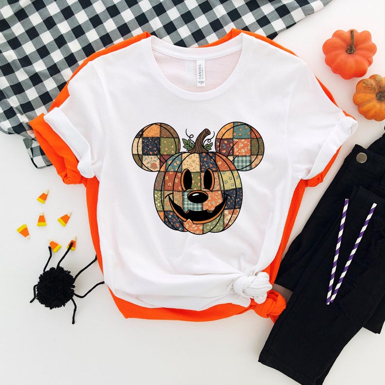 May include: White t-shirt with an orange trim featuring a patchwork pumpkin design in the shape of a mouse. The pumpkin has a smiling face and is surrounded by black pants, candy corn, and a black spider.