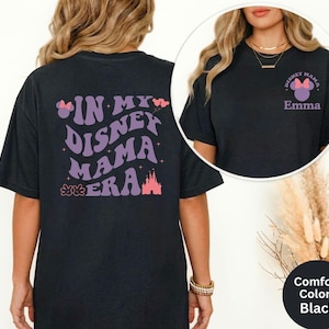 Puede incluir: Camiseta negra con el texto "IN MY DISNEY MAMA ERA" en morado, junto con un castillo y un lazo de Minnie Mouse. La parte delantera de la camiseta tiene un peque&ntilde;o gr&aacute;fico que dice "Disney Mama" y el nombre "Emma".