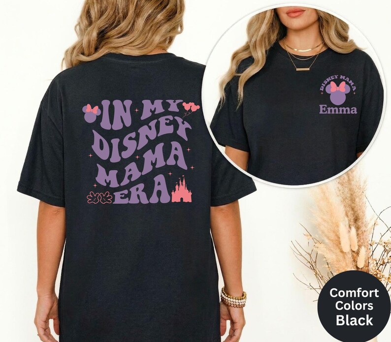 Puede incluir: Camiseta negra con el texto "IN MY DISNEY MAMA ERA" en morado, junto con un castillo y un lazo de Minnie Mouse. La parte delantera de la camiseta tiene un peque&ntilde;o gr&aacute;fico que dice "Disney Mama" y el nombre "Emma".