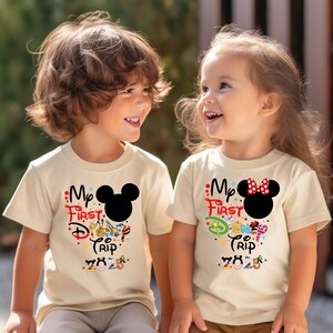 Peut inclure: Deux enfants portant des t-shirts cr&egrave;me avec l'inscription "My First Disney Trip 2025". L'un des t-shirts pr&eacute;sente une t&ecirc;te de Mickey Mouse noire, l'autre une t&ecirc;te de Minnie Mouse avec un n&oelig;ud rouge.