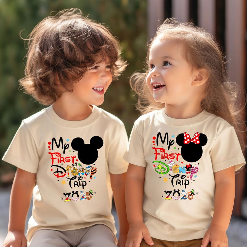 Peut inclure: Deux enfants portant des t-shirts cr&egrave;me avec l'inscription "My First Disney Trip 2025". L'un des t-shirts pr&eacute;sente une t&ecirc;te de Mickey Mouse noire, l'autre une t&ecirc;te de Minnie Mouse avec un n&oelig;ud rouge.