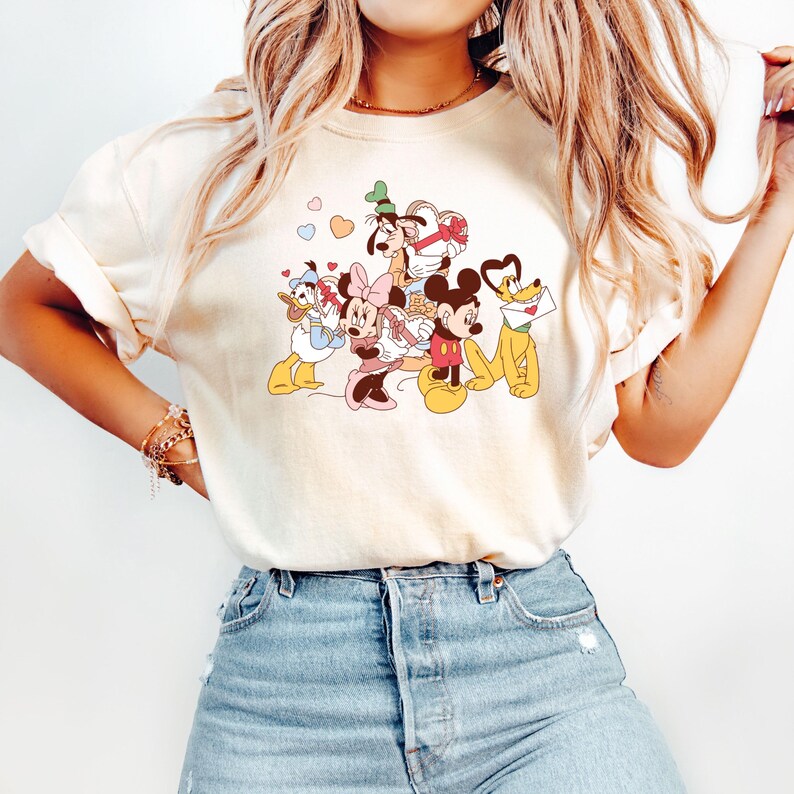 May include: A white t-shirt with a graphic print of Mickey Mouse, Minnie Mouse, Donald Duck, Goofy, and Pluto. The characters are surrounded by hearts and are in a playful pose.