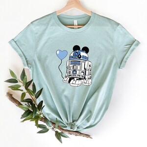 May include: A light blue t-shirt featuring a graphic of R2-D2 with Mickey Mouse ears holding a blue heart-shaped balloon. The design is in white, blue, and black. The shirt is displayed on a wooden hanger.