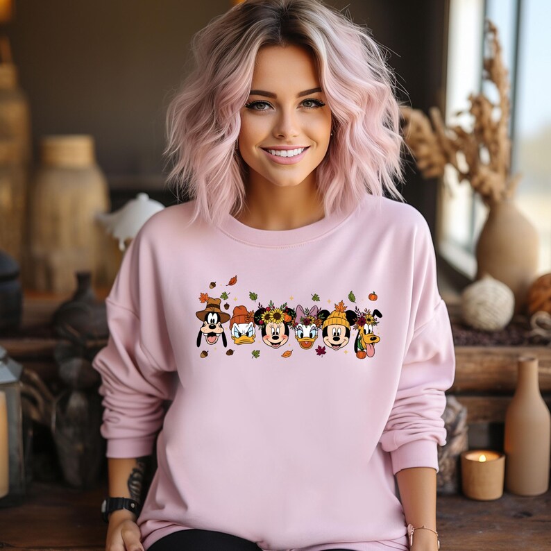 May include: A light pink sweatshirt featuring a graphic of Disney characters in fall attire. The characters include Mickey Mouse, Donald Duck, Daisy Duck, Minnie Mouse, and Pluto, with autumn leaves and pumpkins. The sweatshirt has long sleeves.