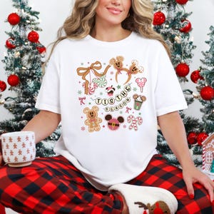May include: White t-shirt with a festive design featuring gingerbread men, Mickey Mouse ears, candy canes, and the phrase "Tis the Season." The design includes pink, brown, and green accents, perfect for the holiday season.