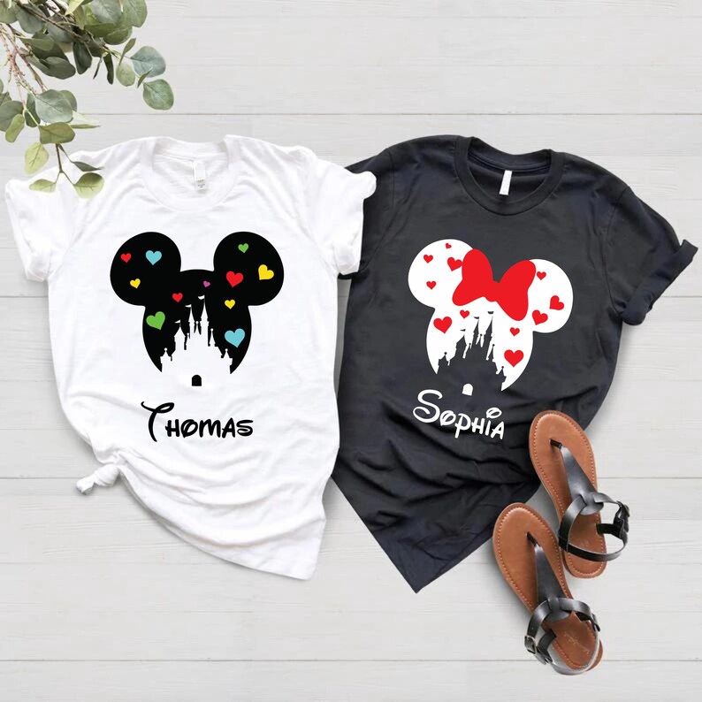 May include: Two white t-shirts with a black Mickey Mouse silhouette and a black castle with colourful hearts. The text "Thomas" is printed on one shirt. Two black t-shirts with a white Minnie Mouse silhouette and a white castle with red hearts. The text "Sophia" is printed on one shirt.