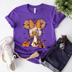 May include: Purple t-shirt with a graphic of a cartoon character with a bow and a castle in the background. The character is holding a pumpkin and there are bats and pumpkins around the castle.