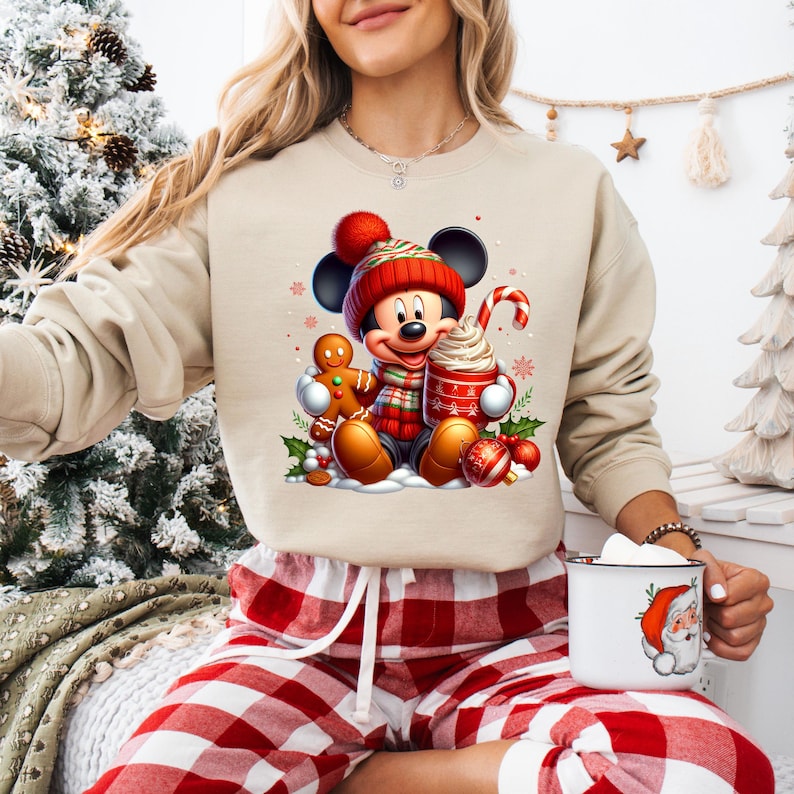 May include: A beige sweatshirt with a Christmas-themed graphic featuring Mickey Mouse wearing a red hat and scarf, holding a gingerbread man and a cup of hot chocolate. The graphic also includes holly, ornaments, and a candy cane.