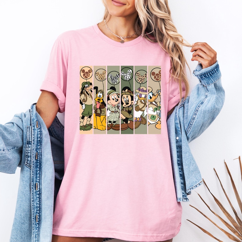 May include: Pink t-shirt with a graphic of Mickey Mouse, Minnie Mouse, Donald Duck, Goofy, and Pluto dressed in safari outfits. The characters are holding balloons with animal print designs.