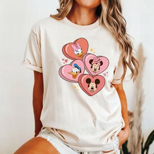 Puede incluir: Una camiseta beige claro con un estampado gr&aacute;fico de Mickey Mouse, Minnie Mouse, Donald Duck y Daisy Duck en corazones rosas.