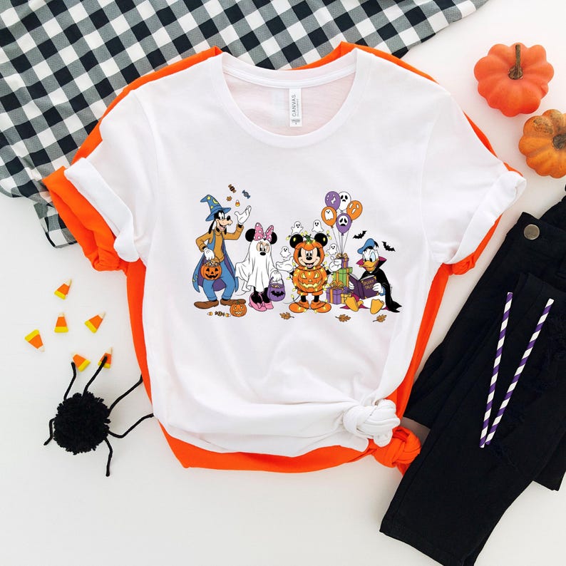 May include: White t-shirt with orange trim featuring a Halloween-themed graphic of Disney characters in costumes. The characters include Goofy, Minnie Mouse, Mickey Mouse, Donald Duck, and others. The shirt is knotted at the bottom.