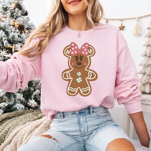 May include: A pink sweatshirt featuring a gingerbread person design. The gingerbread person is wearing a pink bow with white polka dots and has a green candy button on its chest.