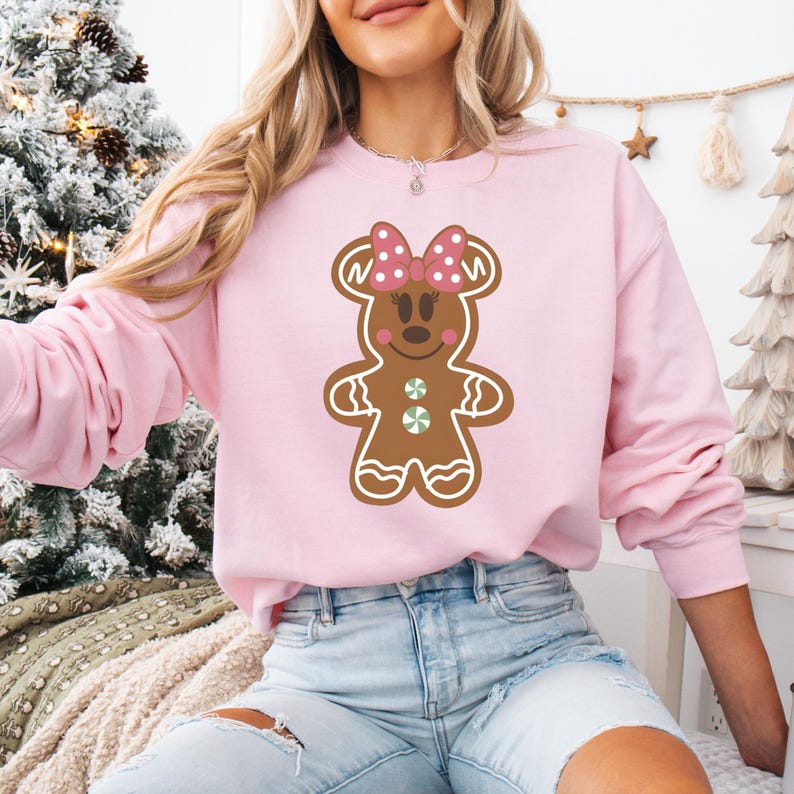 May include: A pink sweatshirt featuring a gingerbread person design. The gingerbread person is wearing a pink bow with white polka dots and has a green candy button on its chest.