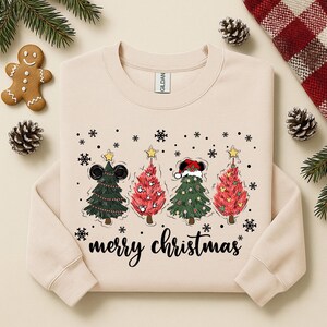 May include: Beige sweatshirt with a festive design featuring four Christmas trees with Mickey Mouse ears and Santa hats. The words "merry christmas" are written in a script font below the trees. Snowflakes and stars complete the holiday theme.