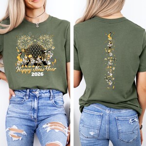 May include: Olive green t-shirt with a festive design for the New Year. The front features fireworks, a geometric sphere, and cartoon characters with the text "Happy New Year 2026." The back has a vertical line of cartoon characters. The model is wearing blue jeans.