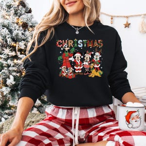 May include: Black Christmas sweatshirt featuring Mickey Mouse, Minnie Mouse, and friends in festive outfits. The word "CHRISTMAS" is written across the top in colorful letters. The sweatshirt is paired with red and white plaid pajama pants.