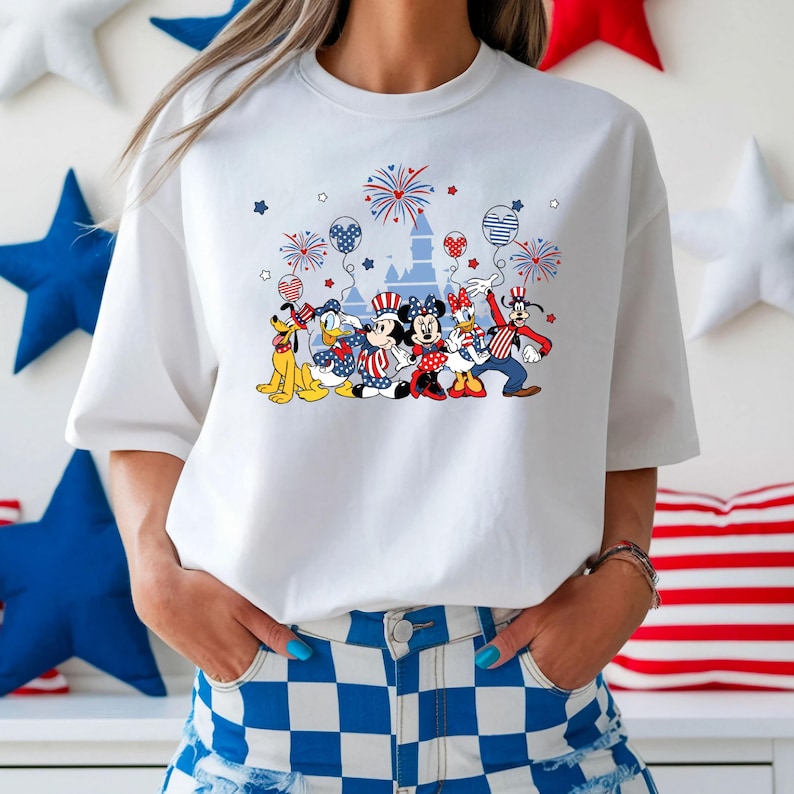 K&ouml;nnte beinhalten: Wei&szlig;es T-Shirt mit einem patriotischen Disney-Design. Die Grafik zeigt Micky und Minnie Maus, Donald Duck und Goofy, zusammen mit Feuerwerk, Luftballons und einem Schloss, in Rot, Wei&szlig; und Blau. Das T-Shirt ist ein l&auml;ssiger Stil.