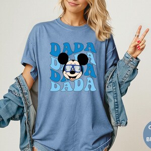 K&ouml;nnte beinhalten: Blaues T-Shirt mit einer Grafik von Mickey Mouse mit Brille und dem Wort "DADA" in einem Retro-Schriftzug. Das Shirt ist im Comfort-Colors-Stil in Jeansblau.