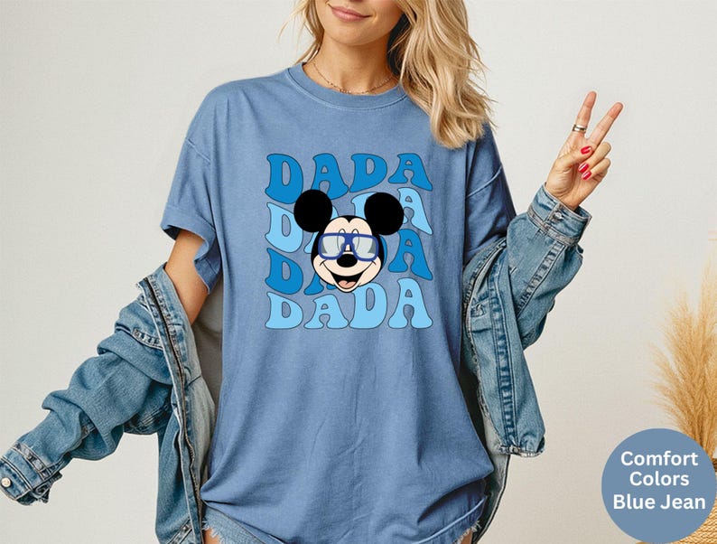 K&ouml;nnte beinhalten: Blaues T-Shirt mit einer Grafik von Mickey Mouse mit Brille und dem Wort "DADA" in einem Retro-Schriftzug. Das Shirt ist im Comfort-Colors-Stil in Jeansblau.