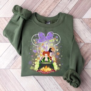 May include: Green sweatshirt with a graphic of the Sanderson sisters from Hocus Pocus in a Mickey Mouse silhouette. The graphic includes a cauldron, a castle, and the text "Hocus Pocus".