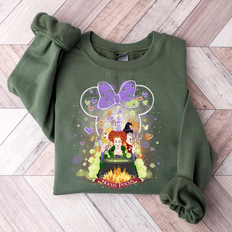 May include: Green sweatshirt with a graphic of the Sanderson sisters from Hocus Pocus in a Mickey Mouse silhouette. The graphic includes a cauldron, a castle, and the text "Hocus Pocus".