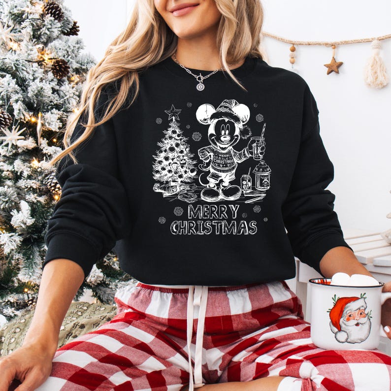 May include: Black sweatshirt featuring a white graphic of Mickey Mouse holding a lantern and standing next to a Christmas tree. The text "MERRY CHRISTMAS" is below the graphic. The person is wearing red and white plaid pajama pants.