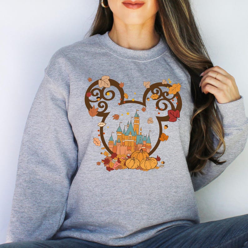 May include: A gray sweatshirt with a graphic of Mickey Mouse ears with a fall-themed design. The design features a castle, pumpkins, and leaves in shades of orange, brown, and yellow.