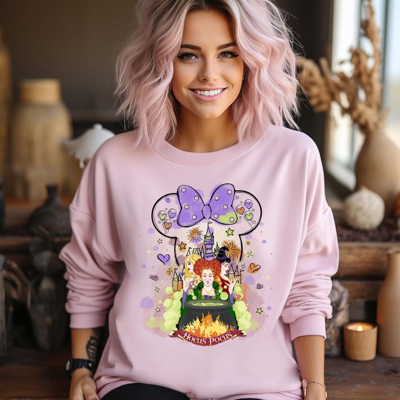 May include: Pink sweatshirt with a purple and white Mickey Mouse head design featuring the Sanderson sisters from Hocus Pocus. The design includes a cauldron, a castle, and fireworks. The text "Hocus Pocus" is written below the design.