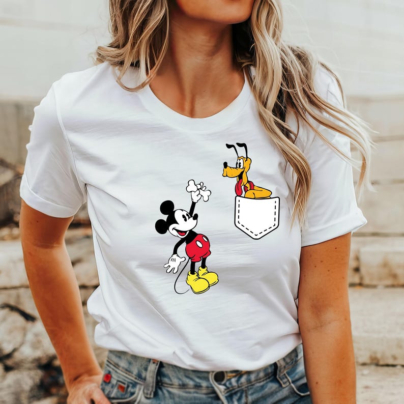 Puede incluir: Camiseta blanca con un gr&aacute;fico de Mickey Mouse sosteniendo un hueso y Pluto asom&aacute;ndose de un bolsillo.