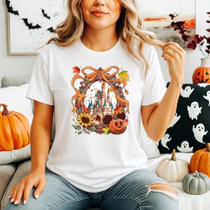 May include: White t-shirt featuring a colorful autumn-themed graphic. The design includes a castle, Mickey Mouse ears, a jack-o'-lantern, sunflowers, and fall leaves. The shirt is a short-sleeved crew neck.