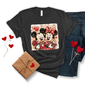May include: Dark gray t-shirt featuring a graphic of Mickey and Minnie Mouse in formal attire, holding roses and chocolates, with heart-shaped balloons. The shirt is paired with blue jeans and a wrapped gift with a heart.