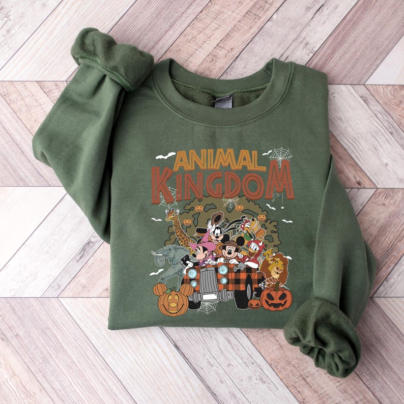Op de afbeelding: Een olijfgroene sweatshirt met de woorden "ANIMAL KINGDOM" in oranje en bruin. De afbeelding toont stripfiguren in een jeep met pompoenen en spinnenwebben, wat een Halloween-thema cre&euml;ert.