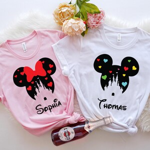 May include: Two white t-shirts featuring a black silhouette of Mickey Mouse ears with a castle inside and colourful hearts. The shirts have the names "Sophia" and "Thomas" printed on them.