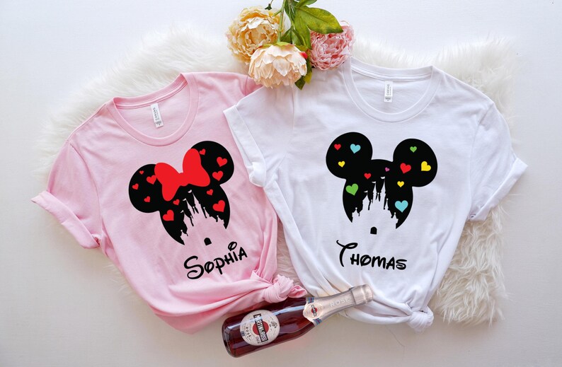 May include: Two white t-shirts featuring a black silhouette of Mickey Mouse ears with a castle inside and colourful hearts. The shirts have the names "Sophia" and "Thomas" printed on them.