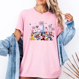 K&ouml;nnte beinhalten: Hellrosa T-Shirt mit einem Aufdruck von Disney-Figuren in patriotischer Kleidung, darunter Micky und Minnie Maus, Donald Duck und Goofy, mit Feuerwerk und einem Schloss. Das Design verwendet die Farben Rot, Wei&szlig; und Blau.