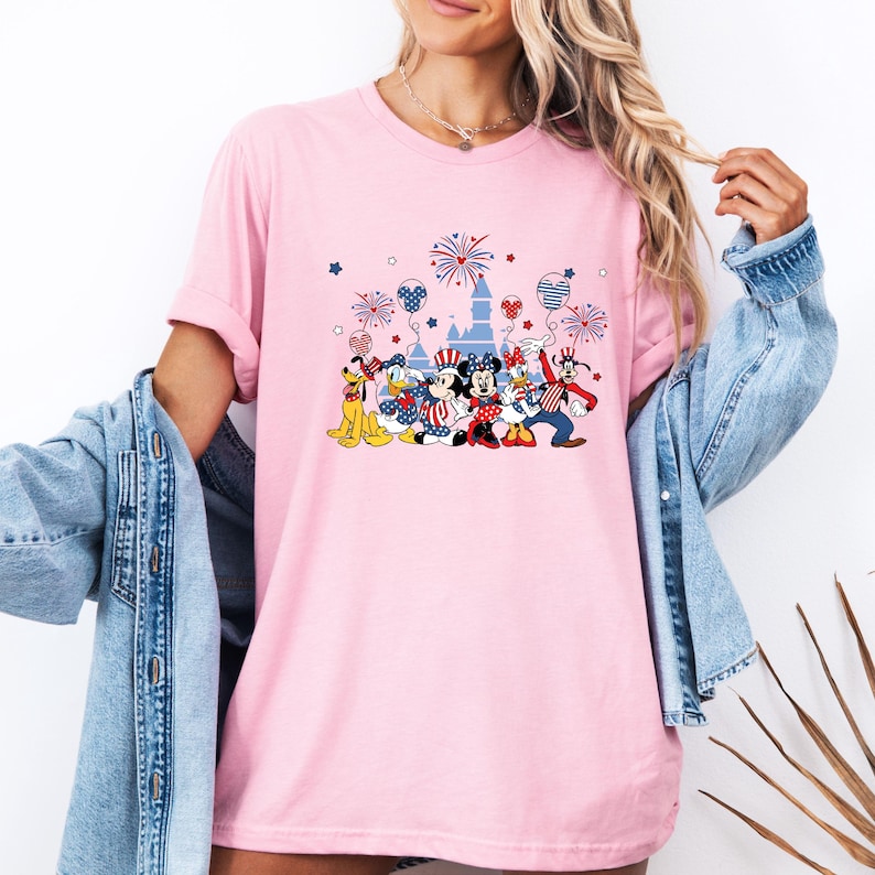 K&ouml;nnte beinhalten: Hellrosa T-Shirt mit einem Aufdruck von Disney-Figuren in patriotischer Kleidung, darunter Micky und Minnie Maus, Donald Duck und Goofy, mit Feuerwerk und einem Schloss. Das Design verwendet die Farben Rot, Wei&szlig; und Blau.