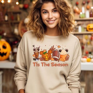 K&ouml;nnte beinhalten: Ein beiger Pullover mit einem Aufdruck einer K&uuml;rbis, einem Football und einer Kaffeetasse mit Mickey Mouse Ohren. Der Text "Tis The Season" ist unter dem Aufdruck gedruckt.