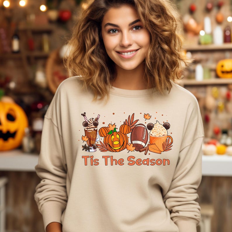K&ouml;nnte beinhalten: Ein beiger Pullover mit einem Aufdruck einer K&uuml;rbis, einem Football und einer Kaffeetasse mit Mickey Mouse Ohren. Der Text "Tis The Season" ist unter dem Aufdruck gedruckt.