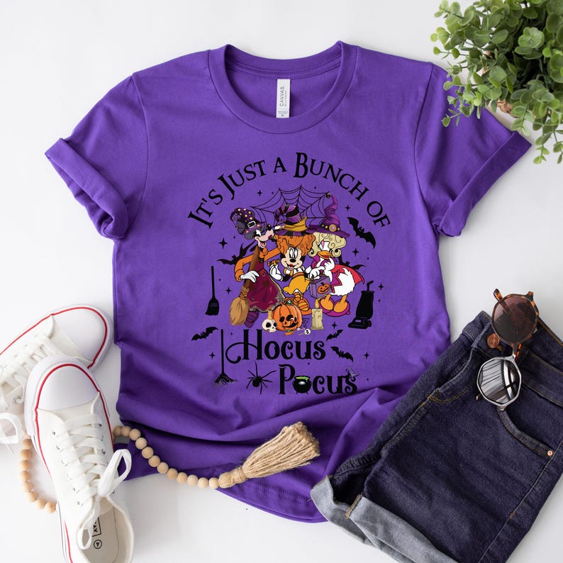 May include: Purple t-shirt with a graphic of Mickey Mouse, Minnie Mouse, Donald Duck, and Goofy dressed as witches. The text "It's Just A Bunch Of Hocus Pocus" is printed on the shirt.
