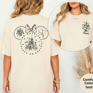 May include: Ivory-colored t-shirt featuring a black graphic of Mickey Mouse ears with a bow, fireworks, and balloons. The text reads "THE HAPPIEST PLACE ON EARTH." A smaller graphic on the front reads "Mommy."