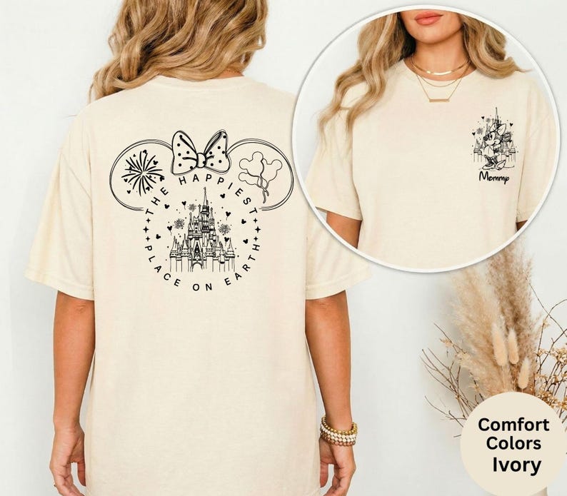 May include: Ivory-colored t-shirt featuring a black graphic of Mickey Mouse ears with a bow, fireworks, and balloons. The text reads "THE HAPPIEST PLACE ON EARTH." A smaller graphic on the front reads "Mommy."