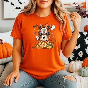 May include: Orange short-sleeved t-shirt with a Halloween graphic. The design shows Mickey and Minnie Mouse atop a carved pumpkin, with ghosts, bats, and sweets. The shirt is made of a soft material.