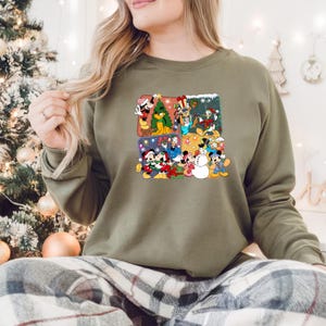 May include: Olive green sweatshirt featuring a colorful graphic of Disney characters celebrating Christmas. The graphic includes Mickey Mouse, Minnie Mouse, Donald Duck, and other characters in festive scenes with a Christmas tree and snow.